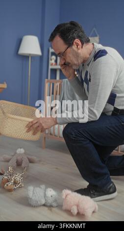 Homme au téléphone plaçant le lapin en peluche dans le panier à côté du berceau dans le bâtiment de la chambre de bébé ; organisation des soins. Banque D'Images