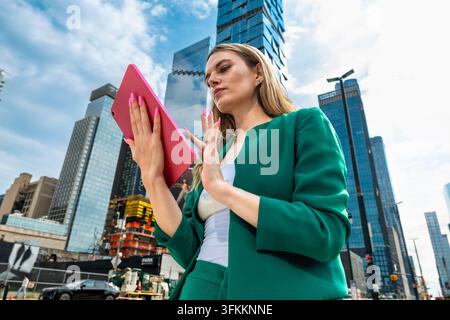 Dirigeante féminine en costume vert se tient à Manhattan avec une tablette rose, gesticulant avec assurance devant des gratte-ciel imposants, symbolisant les affaires modernes Banque D'Images