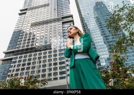 Dirigeante portant un costume vert, debout à l'extérieur, parlant au téléphone portable avec une expression sérieuse, entourée de gratte-ciel modernes dans une ville Banque D'Images