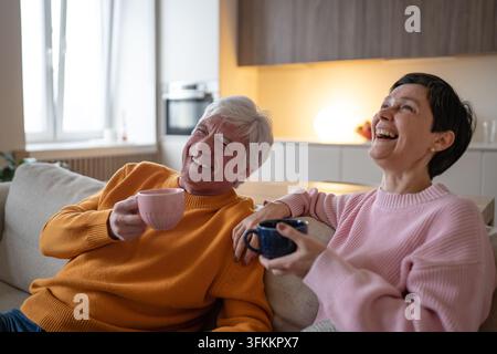 Couple de personnes âgées joyeuses riant ensemble avec des tasses de thé, se relaxant sur le canapé à la maison en retraite Banque D'Images