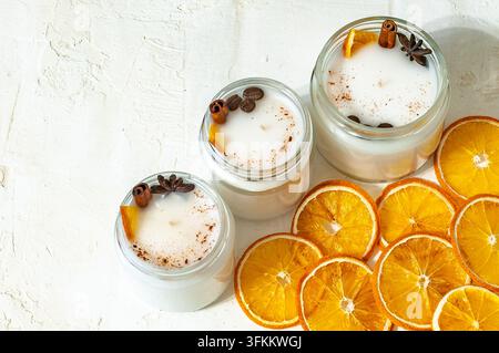 Bougies parfumées dans des bocaux en verre décorés de bâtons de cannelle, d'anis étoilé, de grains de café et de tranches d'orange séchées sur un fond texturé clair. Banque D'Images