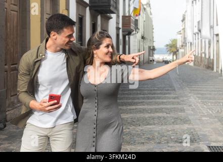 Jeun couple de touristes heureux marchant dans une rue historique de la ville, pointant vers un site emblématique tout en utilisant un smartphone pendant leur visite. Banque D'Images
