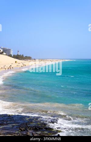 Touristes se détendant sur la plage avec des hôtels et le phare à gauche, Morro Jable, Fuerteventura, îles Canaries. Banque D'Images