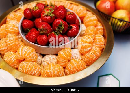 Vue aérienne d'un plateau de service rond doré rempli de fraises rouges fraîches dans un bol central entouré de tranches de mandarine pelées. Photographie alimentaire à angle élevé Banque D'Images