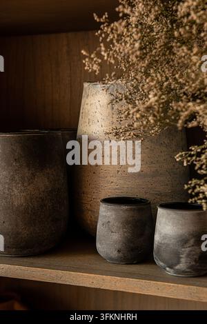 Close-up de tasses en céramique rustique et d'un vase en faïence texturé rempli de délicates fleurs séchées sur une étagère en bois. Banque D'Images
