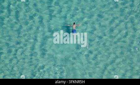 Vue aérienne par le dessus d'un homme nageant sur le dos dans une eau de mer turquoise cristalline à la plage d'Elafonisi sur l'île paradisiaque de Crète, en Grèce. Banque D'Images