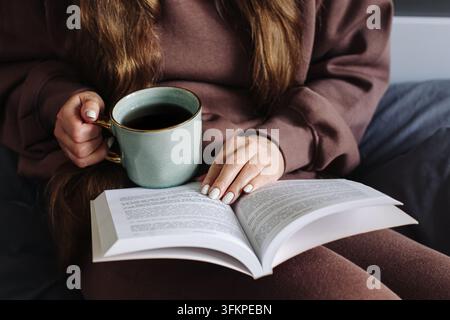 Femme tenant une tasse de thé tout en lisant un livre ouvert. Fond de style de vie confortable pour la détente, boisson chaude, lecture de loisir et moment calme. Banque D'Images