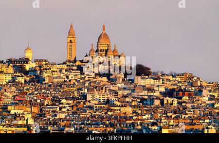 Vue panoramique élevée de la basilique du Sacré-Cœur et de la colline de Montmartre entourée de bâtiments parisiens denses au coucher du soleil à Paris, France. Banque D'Images