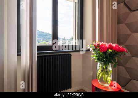 Pivoines et roses roses et rouges dans un vase en verre sur une table d'appoint rouge à côté d'une fenêtre encadrée de noir avec stores beiges dans une pièce conçue à l'italienne. Banque D'Images