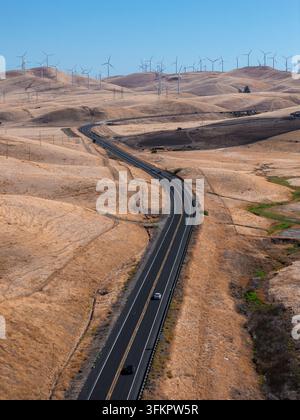 Une vue aérienne montre une autoroute pavée sinuant à travers des collines arides menant vers des éoliennes produisant de l'électricité renouvelable à travers un paysage en Ca Banque D'Images
