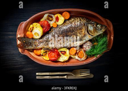 Carpe entière au four avec légumes rôtis, gros plan. Poisson entier délicieux cuit au four servi avec des pommes de terre rôties, des tomates, des carottes et de l'ail dans la rouille Banque D'Images