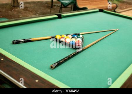 Installation de table de billard d'extérieur avec une toile verte usée et tachée, boules de billard colorées disposées en triangle, mise au point sélective sur les queues dans un parc automnal Banque D'Images