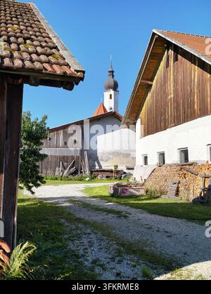 Cour de ferme rurale ensoleillée avec granges en bois, chemin de gravier, toits en tuiles et clocher s'élevant au-dessus des bâtiments rustiques sous un ciel bleu clair, été Banque D'Images