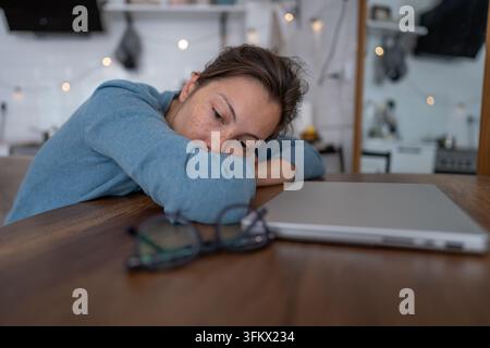 Femme épuisée et débordée assise à une table à côté d'un ordinateur portable fermé après de longues heures de travail sur ordinateur. Banque D'Images