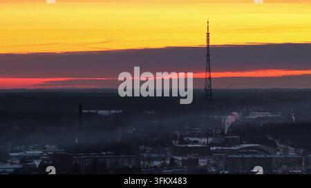 Vue aérienne d'une haute tour radio s'élevant au-dessus d'un paysage urbain brumeux lors d'un coucher de soleil vif avec un ciel orange et rouge sur la Lettonie. Banque D'Images