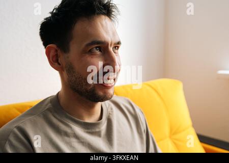 Portrait rapproché d'un homme heureux se détendant à la maison sur un canapé jaune. Expression joyeuse et sourire sincère. Éclairage latéral naturel sur son visage. Chaud, positi Banque D'Images