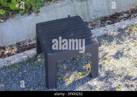 Petite table d'extérieur en plastique noir avec un motif tissé posée sur du gravier à côté d'un canal de drainage en béton. Banque D'Images