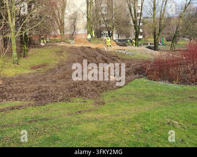 Un chantier de construction dans un cadre de parc urbain présente un grand fossé creusé avec du sol boueux empilé le long de ses bords. Plusieurs ouvriers portant Banque D'Images