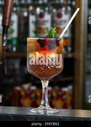 Une boisson riche et ambrée repose dans un verre à cocktail en cristal taillé. Garnie de fruits et d'un brin de menthe, elle semble invitante. Une paille blanche repose dans la boisson, rea Banque D'Images