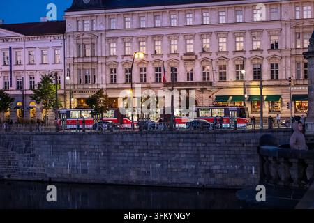 Deux tramways de Prague passent devant des bâtiments historiques au bord du fleuve à la tombée de la nuit sur la Vltava. Prague, République tchèque, 22 septembre 2026. Banque D'Images