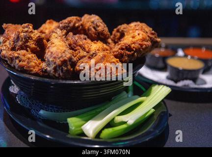 Une photo en gros plan d'un grand bol d'ailes de poulet croustillantes et frites, servies sur une assiette noire avec des tiges de céleri vert frais, accompagnées de sauces de trempage Banque D'Images