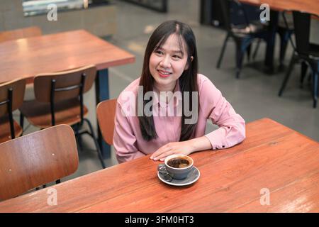 Vu sous un angle élevé, une jeune femme asiatique heureuse en chemise rose est assise à une table en bois dans un café, souriant tout en regardant sur le côté à côté d'une tasse Banque D'Images