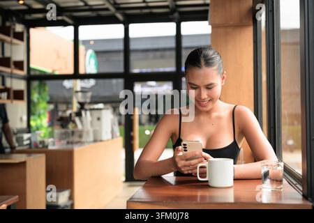 Une photo horizontale authentique et non retouchée d'une jeune femme thaïlandaise heureuse de la région d'Isan dans la vingtaine, souriante tout en utilisant le téléphone à une languette en bois sombre Banque D'Images