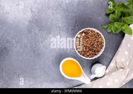 Ingrédients pour un petit-déjeuner sain comprenant de la granola au chocolat, du miel et du lait placés sur un fond en pierre sombre avec des feuilles de menthe fraîche et un napperon à pois Banque D'Images