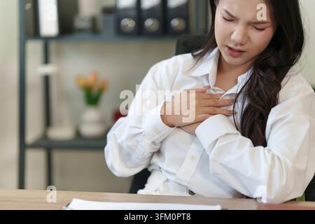 Une jeune femme asiatique se tient la poitrine en proie à la douleur tout en travaillant dans son bureau à domicile, illustrant des symptômes d'anxiété, de stress ou d'un problème de santé plus grave Banque D'Images