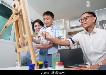 Un plan bas d'un homme asiatique âgé guidant la main de son petit-fils pour peindre sur un chevalet, tandis que le père regarde avec bonheur dans le salon de leur maison Banque D'Images