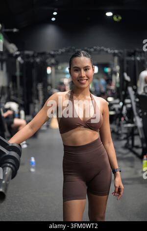 Un jeune influenceur de fitness thaïlandais souriant en vêtements de sport côtelés bruns se tient confiant dans une salle de sport intérieure moderne après une séance d'entraînement réussie, showc Banque D'Images
