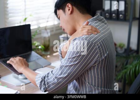 Un homme asiatique ressent des douleurs à l'épaule en travaillant sur son ordinateur portable dans un bureau à domicile, soulignant l'inconfort et le stress liés au travail à distance Banque D'Images