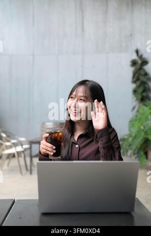 Un plan moyen vertical d'une jeune femme asiatique radieuse souriant largement et agitant la main tout en regardant sur le côté. Elle est assise à une table extérieure derrière un Banque D'Images