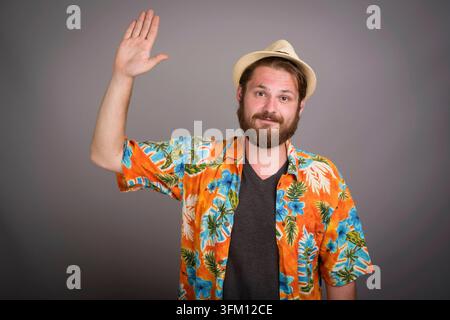 Portrait studio d'un jeune homme barbu dans une chemise tropicale orange colorée et chapeau de paille agitant sa main dans un geste de salutation. Concept d'accueil Banque D'Images