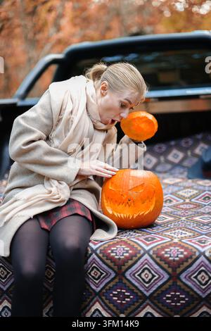 Une femme présente une citrouille jack-o'-lantern faite à la main dans une forêt d'automne, représentant la créativité, les traditions festives et les loisirs en plein air. Banque D'Images