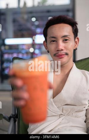Un portrait publicitaire vertical de haute qualité, haute résolution, d'un homme thaïlandais non-binaire gay souriant offrant directement une boisson au spectateur. Le modèle loo Banque D'Images