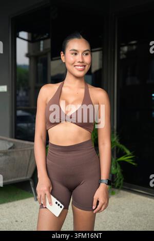 Un jeune influenceur thaïlandais de fitness confiant en vêtements de sport côtelés bruns sourit pour un portrait après une séance d'entraînement réussie dans une salle de sport extérieure Banque D'Images