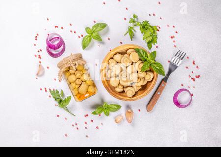 Champignons bien entretenus dans un bocal en verre et un bol en bois entourés de basilic frais, d'anneaux d'oignon rouge, de gousses d'ail, de persil et de grains de poivre rose éparpillés Banque D'Images