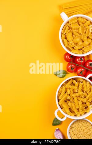 Formes de pâtes crues assorties dans des bols blancs avec tomates fraîches, feuilles de basilic, ail et spaghetti sur un fond jaune vif créant un cul vibrant Banque D'Images