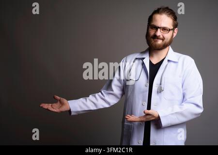 Portrait studio d'un médecin barbu souriant dans une blouse de laboratoire blanche avec un stéthoscope, faisant des gestes avec les mains ouvertes vers l'espace de copie. Hea professionnel Banque D'Images