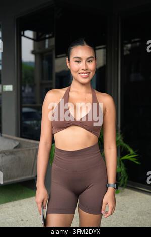 Un jeune influenceur thaïlandais de fitness confiant en vêtements de sport côtelés bruns sourit pour un portrait après une séance d'entraînement réussie dans une salle de sport extérieure Banque D'Images
