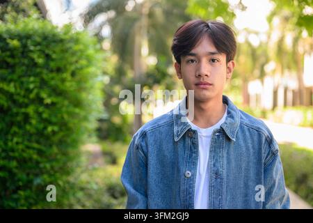 Portrait horizontal moyen d'un beau jeune homme Isan thaïlandais du Sud-est asiatique dans une veste en denim regardant l'appareil photo à l'extérieur avec un backgro bokeh vert Banque D'Images