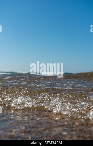Cette photographie capture une vue rapprochée des vagues de l'océan roulant doucement vers le rivage sous un ciel bleu clair et lumineux. L'eau peu profonde révèle int Banque D'Images