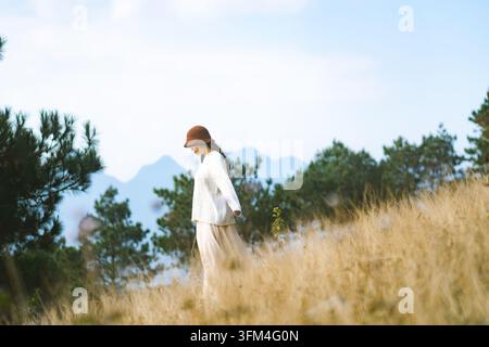 Femme portant un chapeau brun en forme de seau et un pull-over blanc marchant à travers une haute herbe dorée sur une colline montagneuse, Ta Xua, Son La, Vietnam Banque D'Images