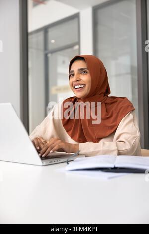 Femme d'affaires heureuse portant des vêtements musulmans assise à son bureau Banque D'Images