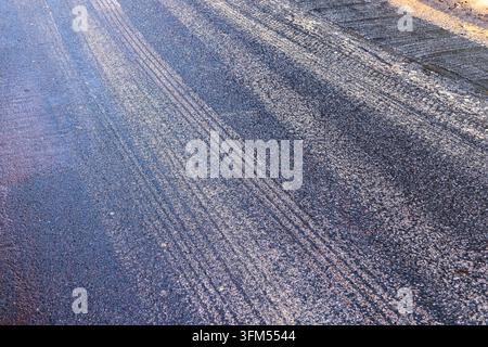 Une surface de route en asphalte rugueuse avec des traces de pneus, une texture de gravier et une lumière chaude du soleil créant un fond urbain abstrait Banque D'Images