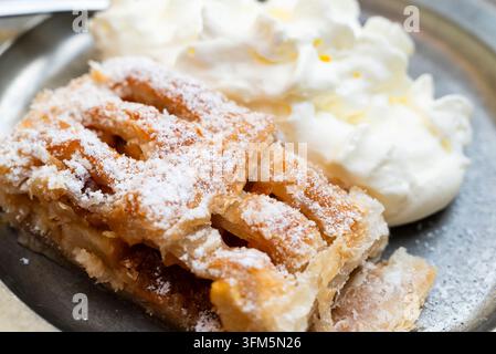 Une tranche d'apfelstrudel allemand chaud saupoudrée de sucre glace et servie avec de la crème fouettée sur une assiette en métal vintage dans un cadre de taverne Banque D'Images