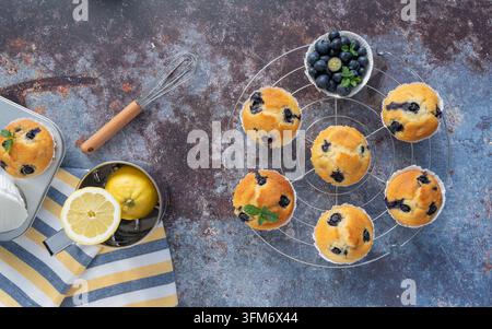 Muffins aux myrtilles faits maison, disposés avec des myrtilles, du citron et des ingrédients de boulangerie sur un fond texturé sombre dans une composition en vue plongeante Banque D'Images
