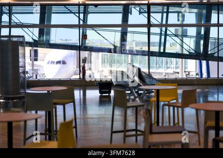 Zone d'attente moderne d'un terminal d'aéroport avec des sièges et des tables vides et un avion de ligne visible à travers de grandes fenêtres au poste d'embarquement Banque D'Images