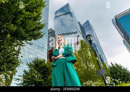 Femme cadre professionnelle en costume vert souriant et téléphonant, debout avec assurance devant des gratte-ciel modernes et des arbres verts à Banque D'Images